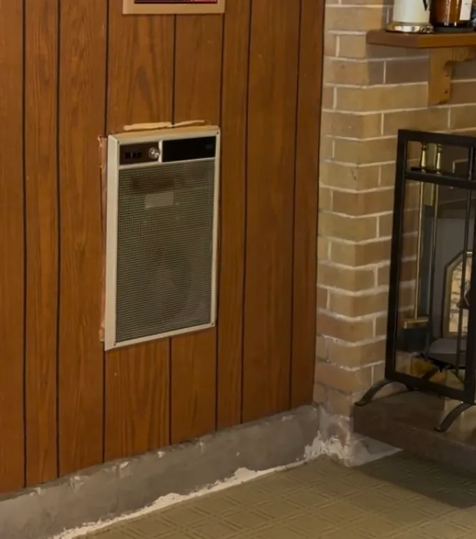 Wall heater set into dark wood paneling beside a brick fireplace