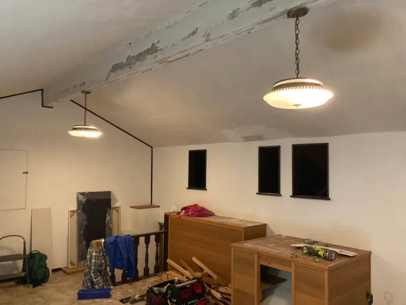 Loft mid-demolition with the old carpet removed, tools and lumber on the floor and a pendant light overhead