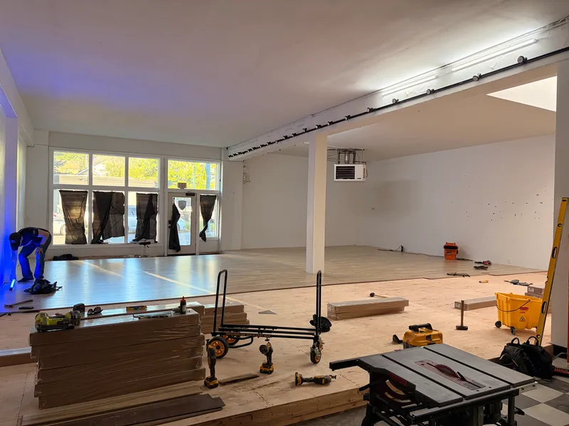 Open room mid-install with a plywood platform, stacks of flooring, a table saw, a cart and a worker kneeling at the far end