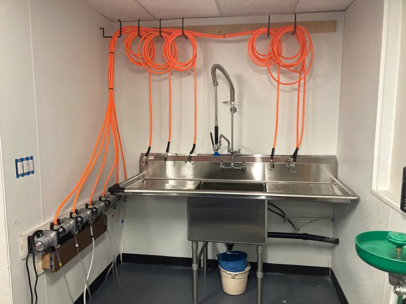 Stainless steel sink with coiled orange hoses hanging above it and a row of fittings along the wall, built as a wash station